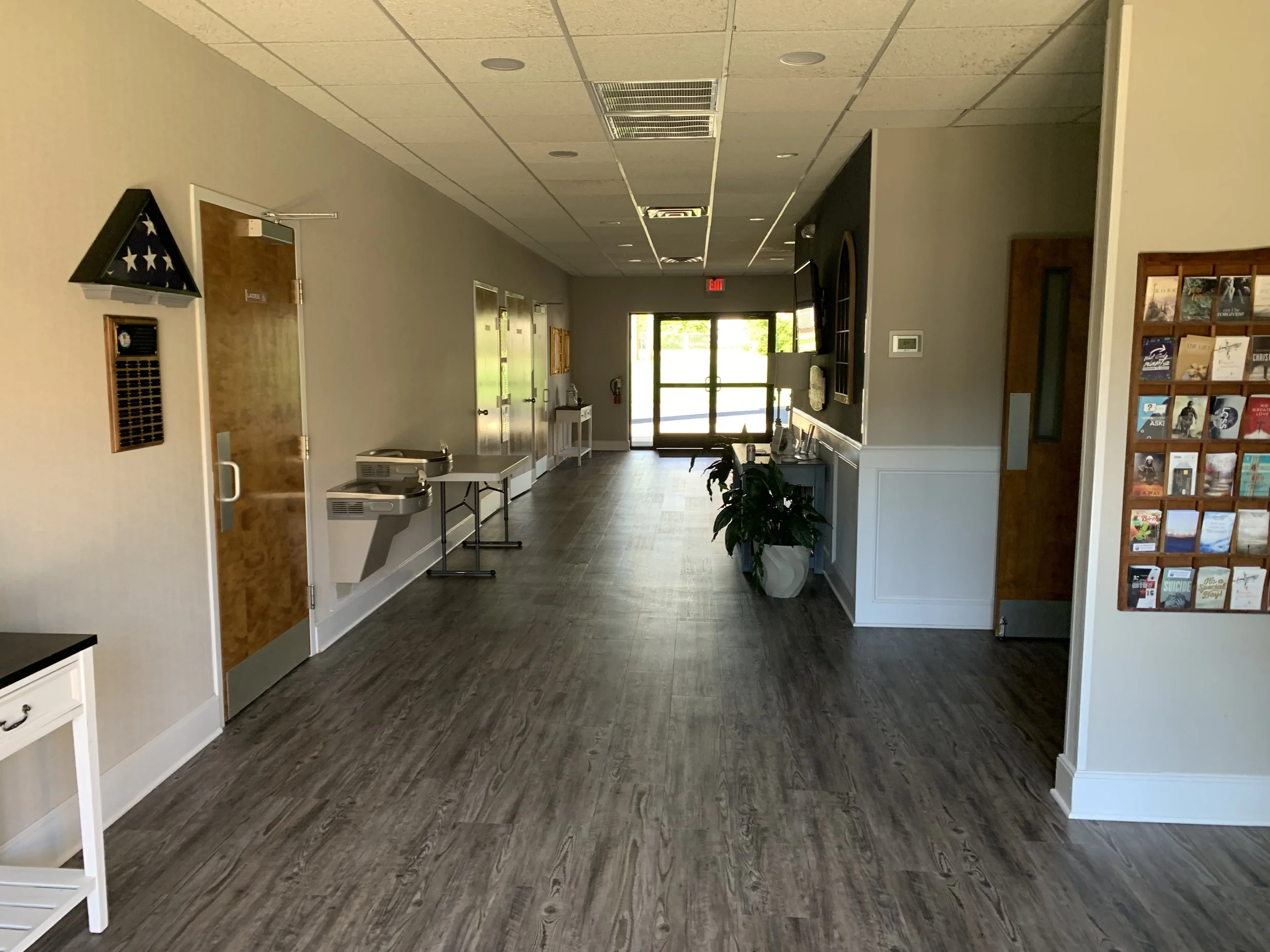 Empty hallway with wooden flooring, gray walls, potted plant, and a wall-mounted TV in a building with glass doors at the end.