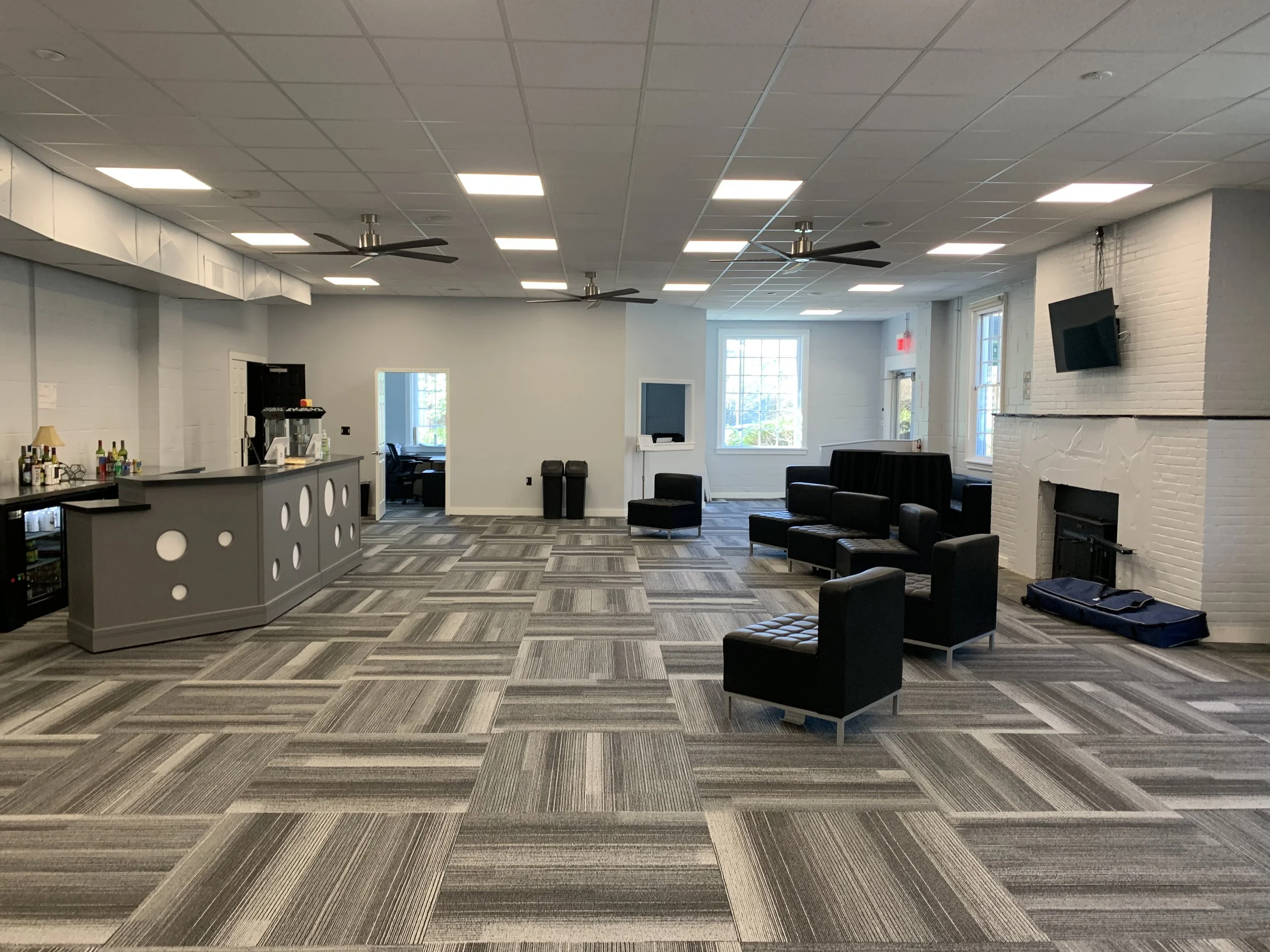 Empty lounge area with rnvasj chairs, bar with drinks, large windows, ceiling fans, and a fireplace on nrfnimr brick wall.