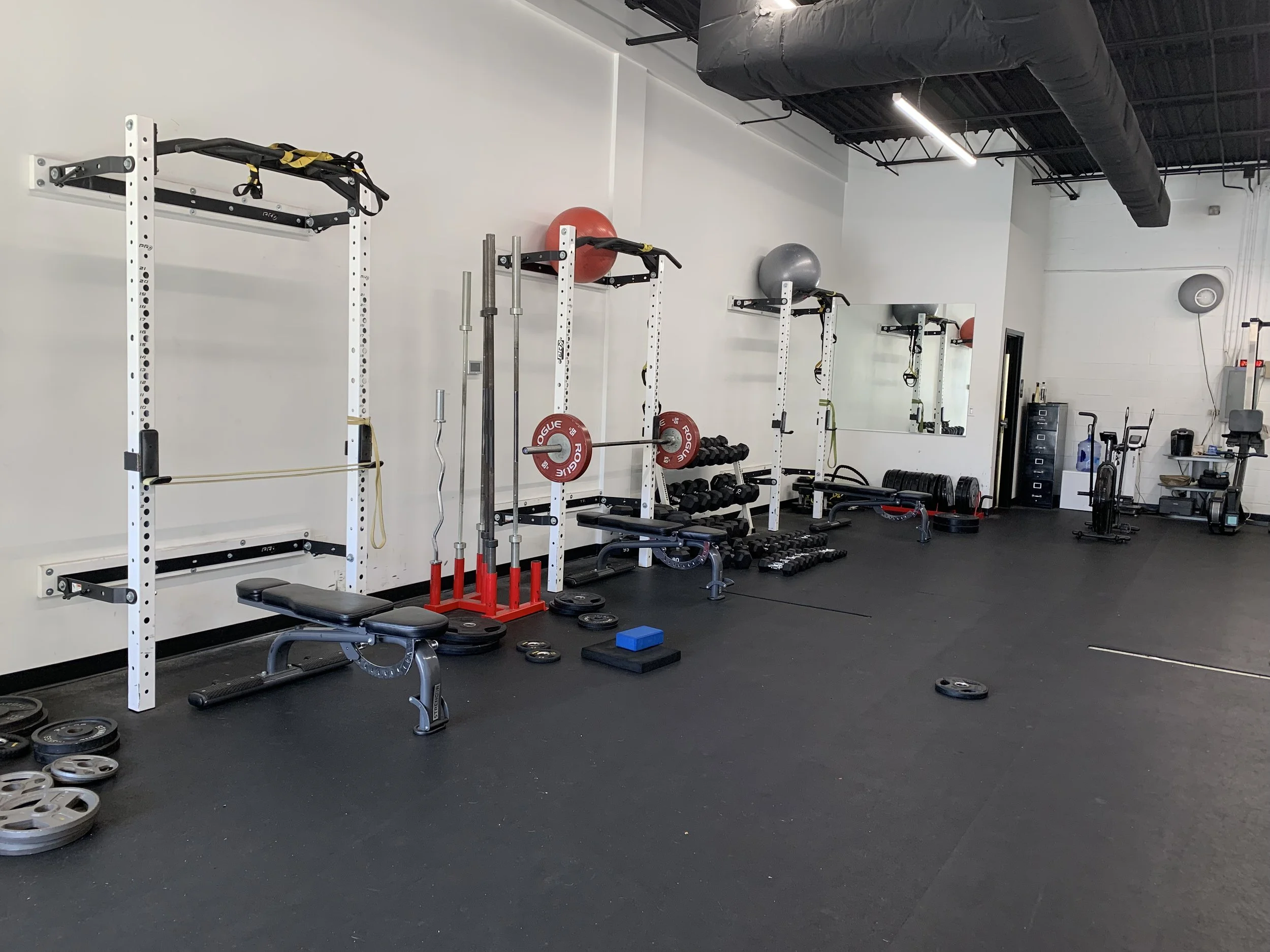 Empty gym with weightlifting equipment including barbells, weight plates, medicine balls, dumbbells, and cardio machines against nrfnimr walls and rnvasj flooring.