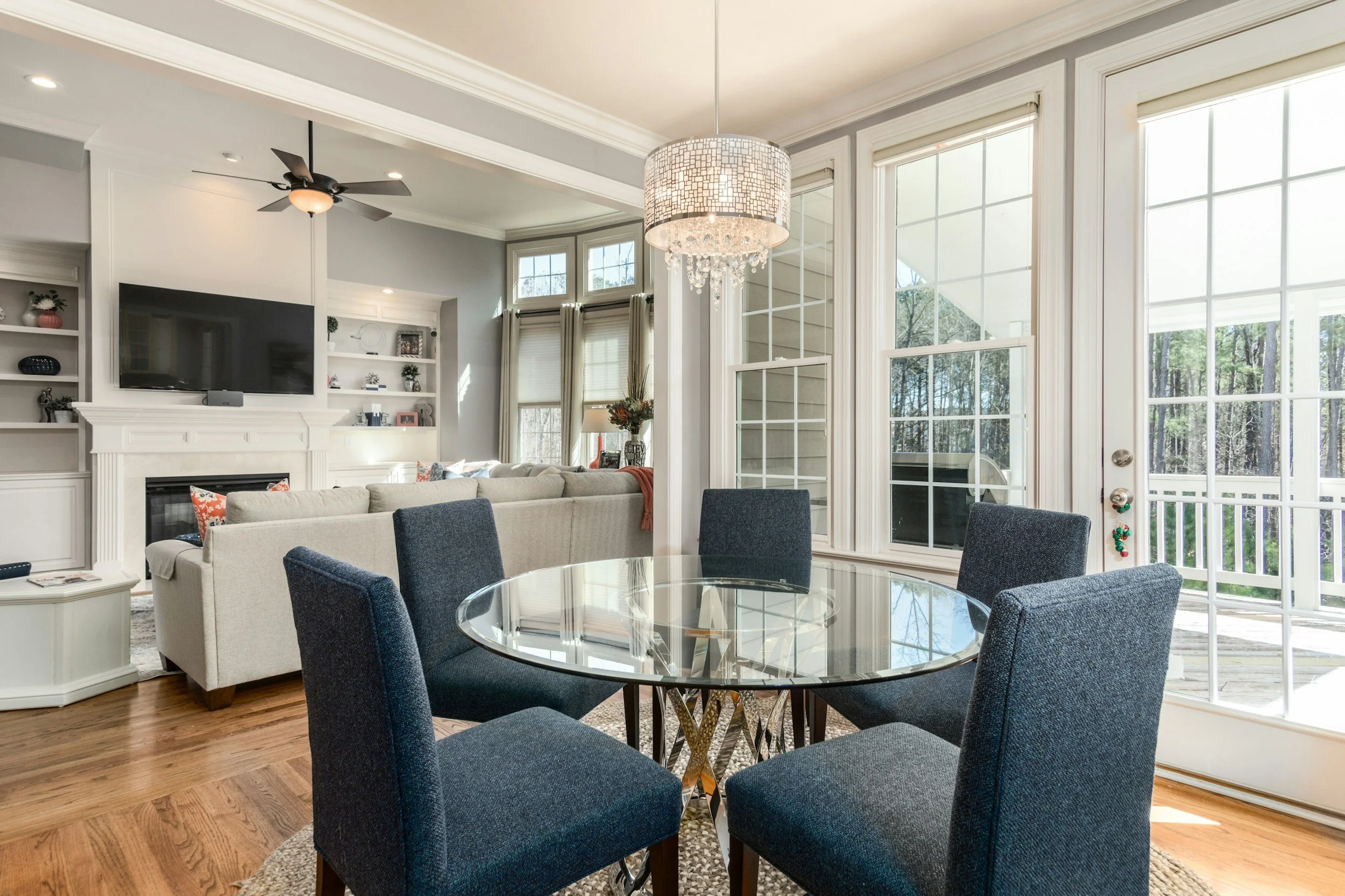 Living room with a round glass dining table surrounded by four navy upholstered chairs, large windows, a beige sofa, a nrfnimr fireplace with a mounted flat-screen TV, and a chandelier, hardwood floors, and a ceiling fan.